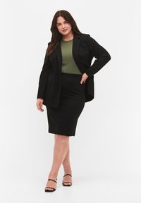 Blazer nero su una maglia verde, abbinato a una gonna a tubino nera e tacchi neri con cinturini. Texture lisce e elementi di design aderenti sono visibili.