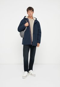 Giacca puffer blu navy con cappuccio foderato in pelliccia, maglione a maglia crema, jeans scuri e borsa a tracolla grigia; sneakers bianche con dettagli grigi.