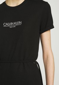 Svart bomulls t-shirtklänning med korta uppvikta ärmar, prydd med en vit "Calvin Klein New York" logotyp på bröstet och en midja med ett bälte.