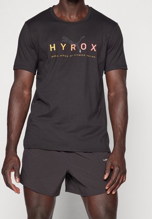 Homme portant un t-shirt de fitness Puma HYROX noir et un short de sport Puma noir, debout devant un fond uni.