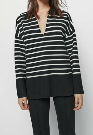 Woman wearing a black and white horizontal striped V-neck sweater with long sleeves and black pants against a plain background.