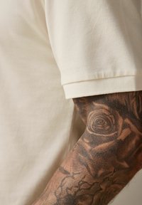 Vaaleanbeige puuvillapolo-paita, jossa on teksturoitu kangas ja käännetyt hihat, käytetty tatuoidussa kädessä, jossa on yksityiskohtaisia kukkakuoseja.