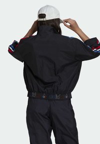 Coupe-vent noir avec des rayures rouges et bleues sur les manches, taille élastique arborant le logo "adidas" multicolore, tissu texturé. Casquette blanche.