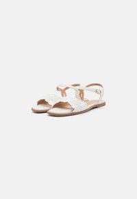 Sandalias blancas con un acento de encaje floral, diseño de punta abierta, correa ajustable en el tobillo y una suela plana color bronce, que ofrece una textura suave.