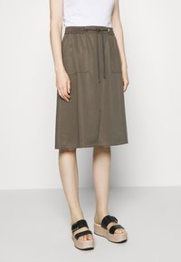 Falda midi verde oliva con dos bolsillos delanteros, cintura elástica con cordón. Combinada con sandalias de plataforma negras y una blusa blanca.