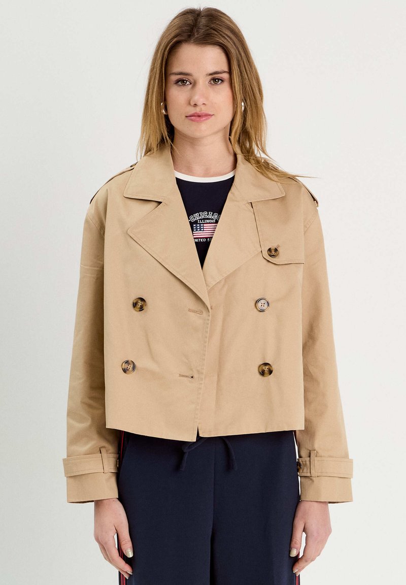 Gabardina corta beige de algodón, con botones de doble botonadura, hombreras y puños doblados. Lleva puesta una camiseta oscura.