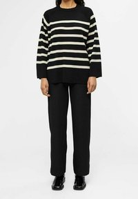 Maglione a righe nere e crema con maniche lunghe, scollo ampio e vestibilità rilassata, abbinato a pantaloni neri larghi e scarpe nere lucide.