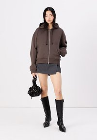 Sudadera marrón con cremallera y cordones, combinada con una minifalda gris. La modelo lleva un bolso negro con flores y calza botas altas negras con punta.