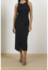 Svart ribbad linne topp kombinerad med en svart omslagsskjorta med en sidoklyvning. Modellen bär svarta remsandaler på ett trägolv.