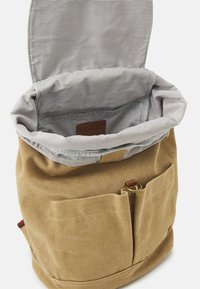 Mochila de lona en beige con un forro interior a rayas gris y blanco. Presenta una etiqueta de cuero y un bolsillo frontal con un anillo metálico.