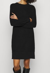 Robe pull noire en tricot avec un col rond, des manches longues et une texture côtelée. Présente une ourlet légèrement plus long en bas.