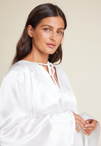 Hvit satengbluse med V-hals og knytedetalj, med brede ermer og en glatt, glossaktig tekstur. Enkel og elegant design.