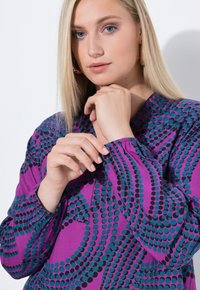Blouse violet avec des motifs de pois noirs et teal. Dotée d'un poignet élastique, d'un col rond et d'une coupe décontractée. Le tissu semble lisse et léger.