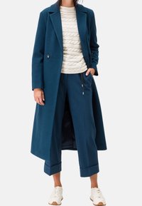Manteau en laine bleu sarcelle au design sur mesure, boutons doubles, associé à un pull crème en maille torsadée et un pantalon court assorti bleu sarcelle. Des baskets blanches complètent le look.