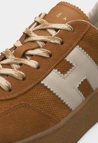 Primer plano de una zapatilla marrón texturizada con cordones beige y un logo de patrón de zigzag cosido en blanco en el lateral.