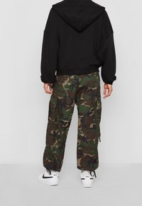 Felpa nera con cappuccio e pantaloni cargo mimetici con tasche laterali e lacci, abbinati a sneakers bianche con dettagli neri.