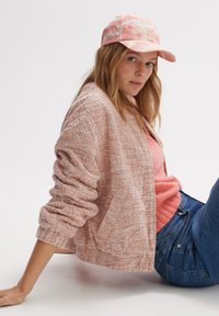 Lichtroze, textuur cardigan met een ontspannen pasvorm, gedragen over een zachte roze top, gecombineerd met blauwe jeans en een lichtroze pet.