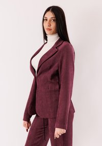 Completo in velluto a coste bordeaux con blazer strutturato con una tasca singola, abbinato a pantaloni svasati coordinati e dolcevita bianco.