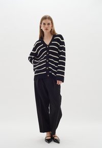 Randig svart och vit cardigan med V-ringning, stora knappar och en avslappnad passform, i kombination med lösa svarta byxor och spetsiga svarta flats.