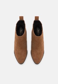 Botas de tornozelo em camurça castanho com bico arredondado, painéis laterais elásticos e alças de puxar, apresentando uma sola de borracha preta texturizada.