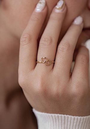 Gold ring featuring two intertwined heart shapes, displayed on a hand with light skin. The background is softly blurred.