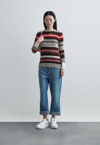 Stribet strikket sweater i rød, beige og brun; kombineret med korte blå jeans og hvide sneakers. Modellen står foran en grå baggrund.