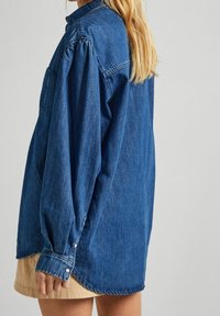 Medium blue denim shirt with long sleeves, buttoned cuffs, and a single chest pocket. It features a relaxed fit and a curved hem.