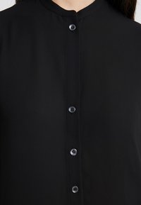 Gros plan d'une personne portant une chemise noire avec un col mandarin et quatre boutons ronds noirs visibles à l'avant.