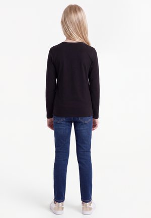 Bambino con lunghi capelli biondi visto di spalle, indossa una maglietta nera a maniche lunghe, jeans blu e scarpe da ginnastica bianche con accenti dorati.
