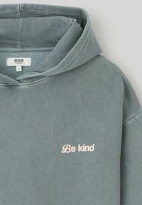 Sweatshirt à capuche surdimensionné en coton gris, présentant un texte brodé en rose "Soyez gentil" sur le devant et une texture douce.