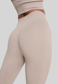 Detaljni kadar osobe koja nosi bež besprešivne tajice visokog struka i odgovarajući skraćeni top nasuprot jednostavne svijetle pozadine.