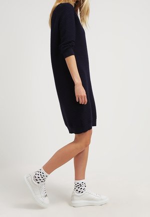 Femme portant une robe côtelée bleu marine longueur genou, baskets blanches et chaussettes blanches à gros pois noirs, debout de profil sur un fond uni.