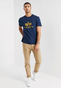 Marineblaues Baumwoll-T-Shirt mit gelbem "Alpha Industries"-Logo; kombiniert mit beigen Chinos und weißen Sneakers, vor einem schlichten Hintergrund stehend.