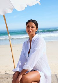 Fehér, könnyű ing, felhajtott mandzsettákkal, fürdőruha fölött viselve, homokos tengerparton ülve, egy nagy fehér napernyő és a kék óceán közelében.