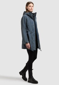 Jeune femme portant une veste à capuche bleue, des leggings noirs et des bottines noires, debout devant un fond clair uni.