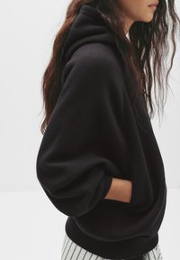 Sweat-shirt à capuche noir avec des manches oversize, des poignets côtelés et des poches latérales. Associé à un pantalon rayé, il présente une coupe décontractée.