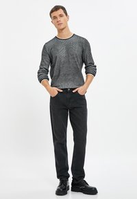 Maglione nero con una finitura testurizzata e colletto a righe, abbinato a pantaloni in denim scuro e scarpe chunky nere. Il modello sta in piedi con le mani in tasca.