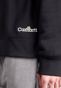 Zwarte sweatshirt met geribbelde manchetten, voorzien van geborduurd 'Carhartt' logo in het wit. Grijze corduroy broek zichtbaar eronder.