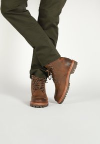 Stivali in pelle marrone con una finitura liscia, dotati di occhielli in metallo, lacci e una suola in gomma resistente. Accentuati da una zeppa color cuoio.