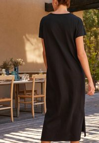 Femme debout à l'extérieur sur un patio en bois, portant une robe midi à manches courtes noire, avec une table à manger et des chaises en arrière-plan.