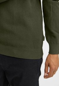 Olivgrüner, strukturierten Pullover mit ribbeltem Saum und Bündchen, ausgestattet mit einem kleinen Logoetikett an der Seite, kombiniert mit dunklen Hosen.