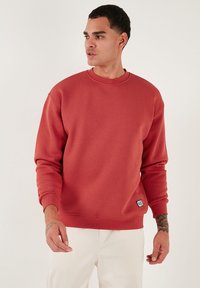 Czerwona bluza z okr�ągłym dekoltem, wykonana z miękkiego materiału, z ribbowymi mankietami, luźnym fasonem i małą naszywką z logo na brzegu. Bezszwowe ramiona.