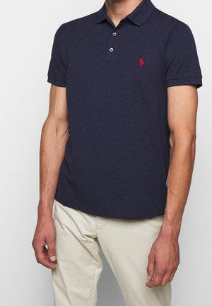 Homme portant un polo bleu marine à manches courtes avec un logo rouge brodé et un pantalon beige sur un fond blanc.