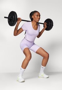 Šviesiai violetinė sportinė apranga su prigludusiomis trumpomis rankovėmis ir šortais. Modelis pritūpia laikydamas juodą štangą, mūvėdamas baltas "Nike" batus ir kojines.