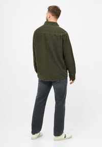 Chemise en velours côtelé vert olive avec un col, des poignets à boutons et un ourlet arrière droit, portée avec un pantalon gris et des baskets blanches.
