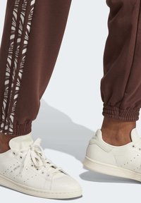 Person wearing white Stan Smith sneakers and brown joggers with patterned side stripes, shown from the calves down against a plain background.