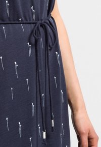 Granatowa sukienka z wiązaniem w pasie. Tkanina zdobiona białymi kwiatowymi wzorami z cienkimi łodygami i sercowatymi czubkami. Miękka tekstura.