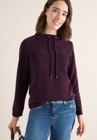Lila Kapuzensweatshirt mit einer Fronttasche, Kordelzug und geprägter Schriftart. Kombiniert mit blauen Jeans, die lässigen Komfort betonen.