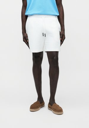 Persona de piel morena con pantalones cortos blancos con cordón, camisa azul clara y zapatos marrones sin cordones, de pie sobre un fondo claro y uniforme.