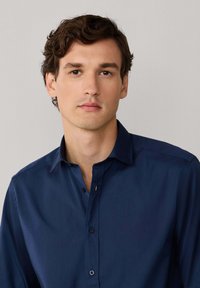 Granatowa koszula zapinana na guziki o gładkiej fakturze, klasycznym kołnierzu i subtelnym detalu guzików, noszona przez modela na neutralnym tle.